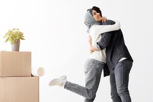 Emotional young muslim lady in hijab hugging her husband, middle-eastern man and woman standing among paper boxes and other stuff in empty room, family moving in new house, full length, copy space photo