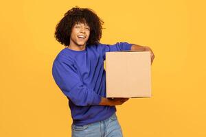 A young black man with curly hair smiles while holding a cardboard box. He stands against a vibrant yellow background, showcasing a cheerful atmosphere perfect for a lifestyle setting. photo