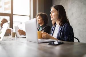A diverse group of professionals is engaged in a meeting at a contemporary office space. They are sharing ideas while working on laptops and enjoying beverages, fostering collaboration and creativity. photo