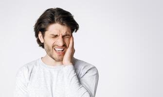 Sharp pain in ears. Man grimaces and holds hand to sore spot isolated on gray background, studio shot photo