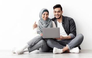 Loving muslim family sitting on floor in new apartment with laptop, buying furniture for house or working on design together, sitting by blank wall, hugging and looking at computer screen, free space photo