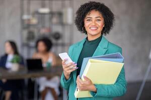 A confident businesswoman stands in a modern office, holding folders and a smartphone. Behind her, colleagues collaborate on a project, emphasizing teamwork and diversity in the workplace. photo