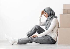 Upset middle-eastern family sitting separated by boxes on floor in empty apartment, having problem with relocating, white wall background, side view, copy space. Frustrated arabic couple at new home photo