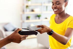 Happy Black Female Customer Paying Courier For Pizza Delivery Putting Smartphone To Payment Terminal Standing In Doorway Of House. Pay Smart With Mobile Banking Application. Selective Focus photo