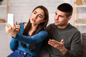 Couple Using Smart Phone For Calling Or Taking Photos, Sitting On Couch In Their Apartment And Gesturing photo