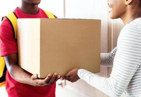 Professional Courier In Uniform Delivering Parcel To African American Lady Customer, Giving Her Paperboard Box Standing In Doorway Of Her Home. Package Delivery Concept. Cropped, Selective Focus photo