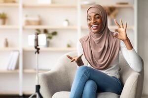Happy Black Islamic Lady In Hijab Using Smartphone On Tripod For Recording At Home, Cheerful Muslim Female Blogger Capturing Content For Her Vlog, Talking And Gesturing At Device Camera photo