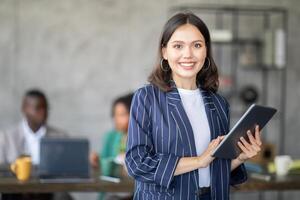 A confident woman stands in front of her diverse team in a contemporary office. She holds a tablet and smiles, as colleagues engage in a productive discussion while working together on projects. photo