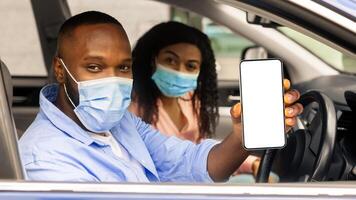 Closeup of black man in surgical mask showing cell phone with white empty screen for mock up sitting on driver seat in car with wife, holding gadget in hand, copy space template. Health passport photo