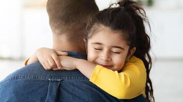 Father's Day Concept. Cute Little Arab Girl Hugging Tight Her Dad At Home, Loving Small Female Child Embracing Daddy With Closed Eyes, Happy Middle Eastern Family Bonding Together In Kitchen, Closeup photo
