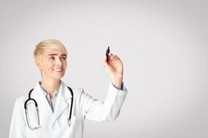 Innovative medicine. Smiling lady adult doctor in white coat with stethoscope using invisible virtual screen, drawing with marker and watch at free space, isolated on gray background, studio shot photo