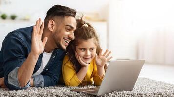 Happy Middle Eastern Father And Female Kid Making Call With Laptop At Home, Arab Dad And Daughter Waving Hands At Web Camera, Communicating With Family Online, Lying On Floor In Kitchen photo
