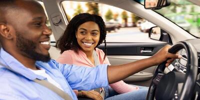 Enjoying Travel Concept. Smiling African American man driving modern luxury car, happy young woman sitting on the front passenger seat, looking on the driver, family going on roadtrip, side view photo
