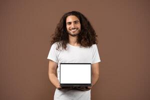 A young European man with long, curly hair stands against a plain brown backdrop, smiling while holding a laptop with a blank screen. He embodies a modern, lifestyle-oriented vibe. photo
