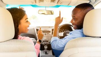 Road Trip Concept. Rear back view of excited playful African American couple driving luxury car, dancing to music and singing favorite song, resting on weekend going on vacation, looking at each other photo