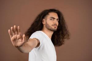 A young European man with long, curly hair stands in a relaxed pose, raising one hand as if signaling to stop. He wears a simple white t-shirt and showcases a thoughtful expression. photo