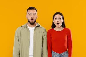 A young couple stands together against a bright yellow background, both displaying expressions of surprise and shock. Their body language reflects a strong bond and shared experience. photo