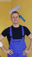 Portrait of a painter man indoors. Young smiling worker in overalls with a painter roller on his head showing yes sign with a thumb. Renovation services. Vertical video
