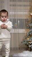 Toddler in white pajama walks by the room. Child holds a pacifier in hands. Christmas tree at backdrop in blur. Vertical video