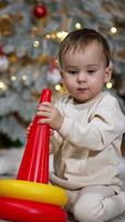 Cute little Caucasian toddler playing with a pyramid sitting on the floor. Christmas tree with a black cat sitting beside at backdrop in blur. Vertical video