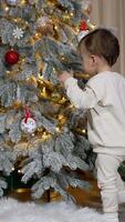 Nice toddler in white clothes wants to take a red ball from Christmas tree. Lovely smiling kid turns around and approaches camera. Vertical video