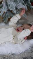 Beautiful toddler lies under Christmas tree waving his little hands. Baby rolls on the soft fluffy plaid trying to stand up. Vertical video