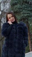 magnifique modèle dans chaud manteau sur la nature Contexte. portrait de une charmant fille dans fourrure manteau parlant le rouge téléphone et à la recherche à caméra dans hiver parc. verticale video