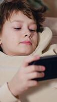 Dark-haired boy lies on couch with phone in hands. Kid plays games and succeeds rejoicing. Christmas tree at backdrop. Vertical video