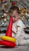 Caucasian toddler sits on the floor playing with a toy pyramid. Decorated Christmas tree at backdrop in blur. Vertical video