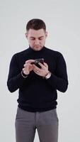 Handsome man with a cell phone isolated in studio. Portrait of a young man in black sweater and grey trousers looking in his phone. Vertical video