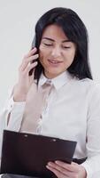 Portrait of a secretary in a blouse talking the mobile phone. Beautiful woman looking into the folder and talking the phone while sitting in studio. Slow motion. Vertical video