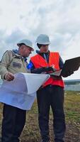 Men in helmets talking about solar panels. Managers discussing the plan on the field with solar panels using a computer laptop. Vertical video