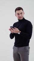 Half-length portrait of man holding a mobile phone. Bearded man in black sweater and grey trousers standing in studio and looking aside with surprise. Slow motion. Vertical video