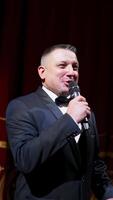 Male announcer with microphone on the stage. Actor in black suit on red scene curtains background. Vertical video