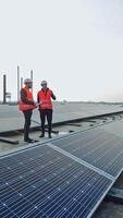 Engineers talk on the solar farm background. Rows of solar panels and two workers in orange uniform and hard hat. Installation of innovative sunny batteries. Vertical video