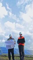 Engineers with laptop and project plan on the solar farm. Workers in special uniform standing among the solar panels and discuss the installation of sunny batteries. Vertical video