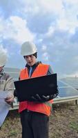 Technicians with plan and laptop on the solar farm. Workers in special uniform discussing the construction of solar panels to get clean energy. Vertical video