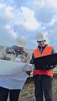 Technicians look into laptop and project plan on the solar farm. Two workers in protective helmets on the background of solar panels. Vertical video