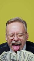 Portrait of happy senior man with money. Mature man in glasses holds many dollar bills and opens his mouth with joyfulness on yellow background. Slow motion. Vertical video