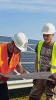 ingenieros hablando en el solar granja. grupo de trabajadores discutir su trabajo mientras mirando a proyecto plan en el soleado baterías antecedentes. vertical video