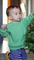 Nice little standing on his knees near Christmas tree. Baby takes a toy from a tree and crawls away. Vertical video