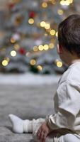Adorable baby sitting his back to camera on the floor looking at Christmas tree. Child quickly crawls to the tree. Vertical video