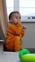 Little kid sitting near the window and the stairs indoors rubs his eyes. Baby pulls the finger into mouth and looks around the room. Vertical video