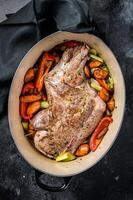 Roasted lamb mutton shoulder meat in a baking dish with vegetables. Black background. Top view photo