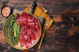 Basturma, sliced dried beef meat, meat Jerky in steel plate with herbs and spices. Wooden background. Top view. Copy space photo