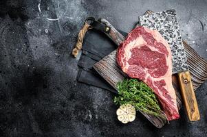 Dry aged Tomahawk rib eye steak, raw beef meat on butcher table. Black background. Top view. Copy space photo