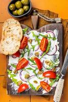 Trendy Butter Board with olives, onion, arugula, capers and tomato. Orange background. Top view photo