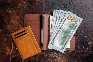 Wooden desk table with blank notebook, pen, wallet with money and credit cards. Brown background. Top view. Copy space photo