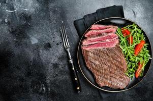Grilled medium rare flank beef steak with salad in a plate. Black background. Top view. Copy space photo