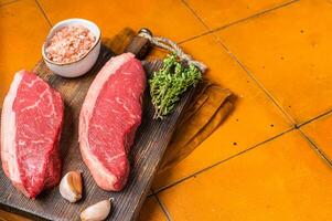 Cap Rump steak, raw beef meat steak with thyme and salt on wooden butcher board. Orange background. Top view. Copy space photo
